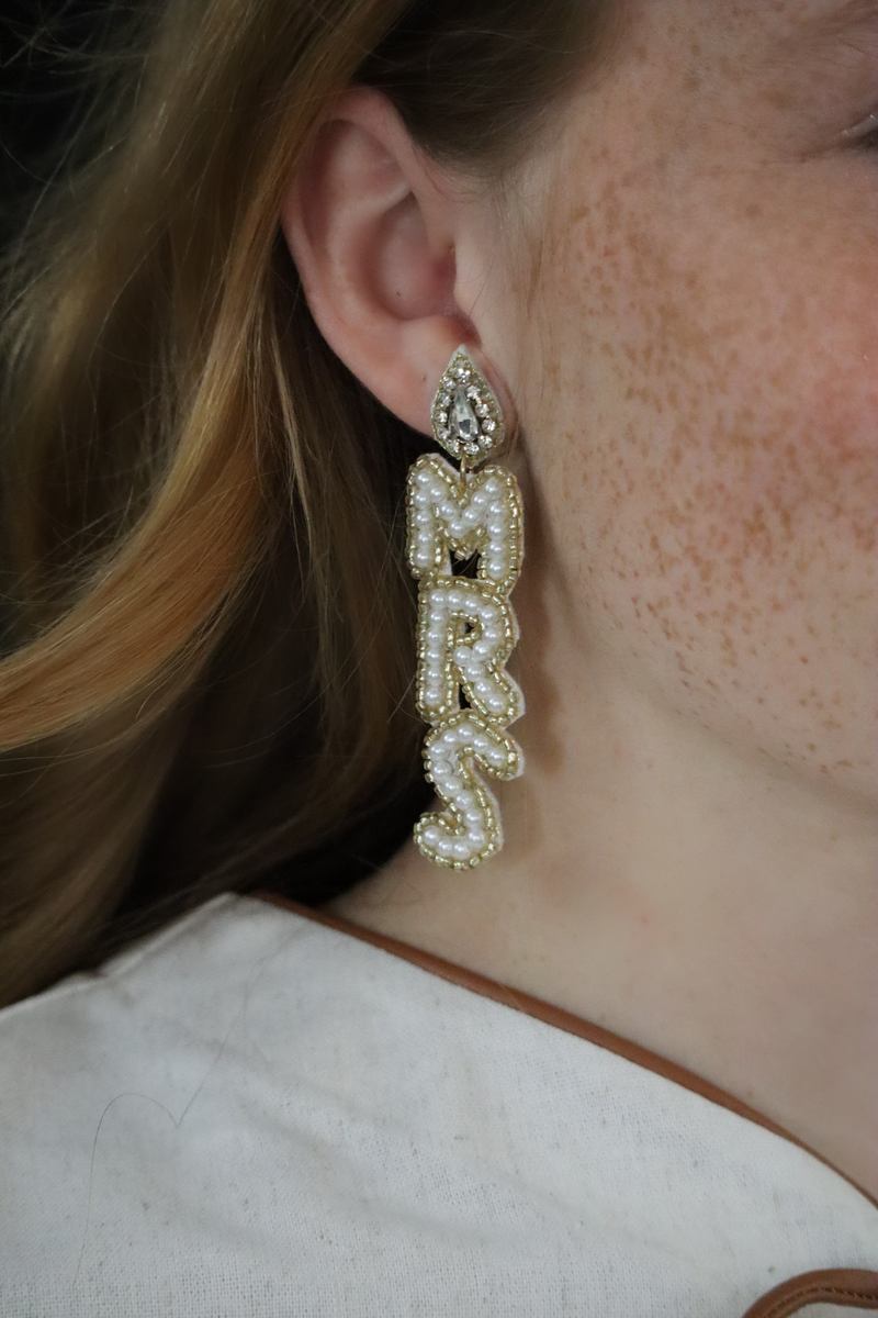 Close-up of a person wearing a 'MRS' earring with pearl details.